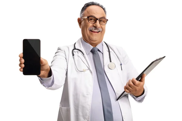 笑顔の成熟した医師は クリップボードを保持し 白い背景に隔離されたスマートフォンを示しています — ストック写真