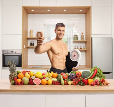 Elinde meyve ve sebze yığını olan üstsüz bir adam, elinde ağırlık terazisi ve mutfak tezgahının arkasında el kol hareketi yapıyor.