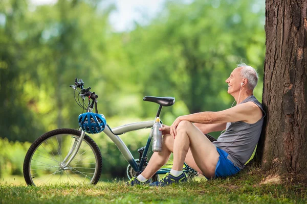 Active senior listening to music seated by a tree in park — Stok fotoğraf