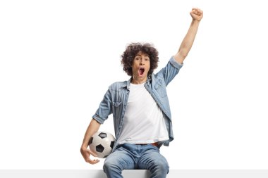 Happy young man cheering and holding a football seated on a panel isolated on white background