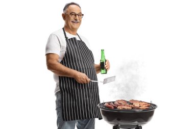 Mature man holding a turner utensil and a bottle of beer and grilling meat on a bbq isolated on white background