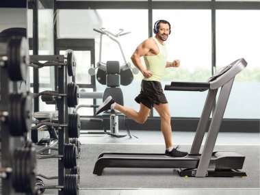 Koşu bandında kulaklıklarıyla koşan bir adamın tam boy çekimi.