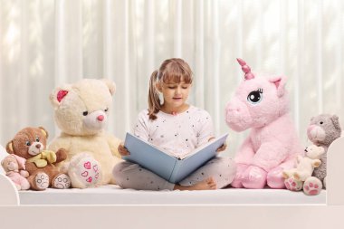 Pijamalı, oyuncaklı, kitap okuyan bir kız.