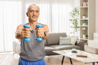 Spor kıyafetli yaşlı bir adam evde halterlerle egzersiz yapıyor.
