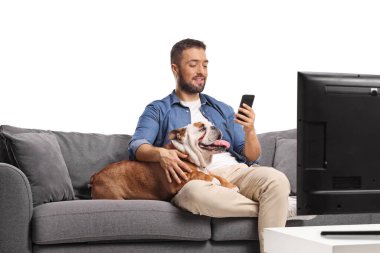 Kanepede köpeğiyle oturan genç bir adam beyaz arka planda izole edilmiş bir akıllı telefon kullanıyor.