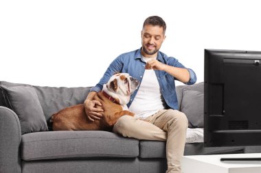 Adam buldoga yemek veriyor ve beyaz arka planda izole bir şekilde televizyon karşısında bir koltukta oturuyor.