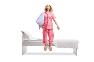 Pijamalı bir kadın yastık tutuyor ve beyaz arka planda izole bir şekilde yatağın önüne atlıyor.