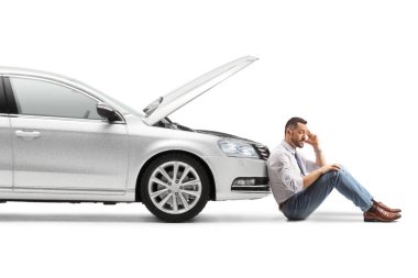 Stresli bir adam kırık bir arabanın yanında oturuyor. Kaputu açık, beyaz tenli.