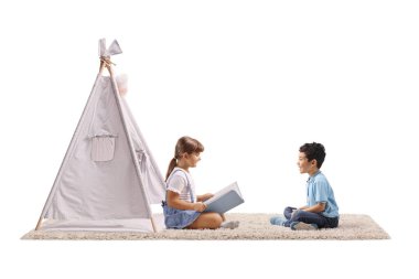 Çocuklar beyaz arka planda izole edilmiş küçük bir çadırın önünde halının üzerinde oturuyorlar.