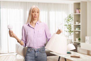 Modern bir oturma odasında elinde ampul ve abajur tutan kafası karışmış bir kadın.