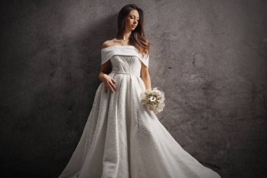 Şık genç gelin elinde bir buket tutuyor ve gri duvara yaslanıyor.