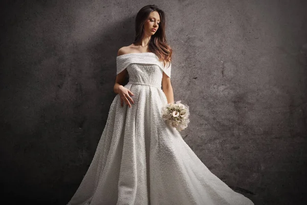 Şık genç gelin elinde bir buket tutuyor ve gri duvara yaslanıyor.