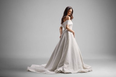 Gri arka planda, omuzları açık gelinlik giyen zarif bir gelinin yan görüntüsü.