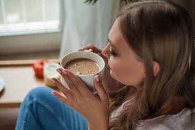 Güzel genç bayan sıcak kakao içiyor ve pencerenin kenarında oturuyor.