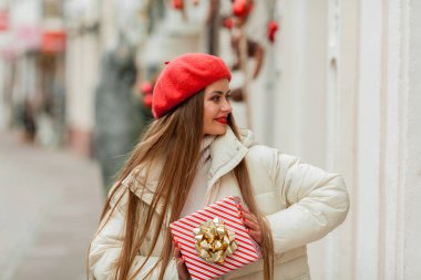 Avrupa 'nın bir şehrinin caddesinde elinde hediye kutusuyla kırmızı bereli genç bir kadının portresi. Noel alışverişi.