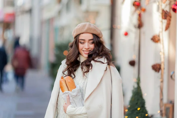Portrait of a young beautiful woman in a beret in a European city. Young woman holds a paper bag with baguettes. Christmas.