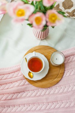 Bir kupa sıcak çay, pembe örgü battaniye, vazoda bir buket lale. Yatakta kahvaltı. Samimi.