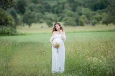 Beyaz elbiseli, kıvırcık uzun saçlı güzel, hamile bir kadın tarlada yürüyor. Hamile bir kadının bahar portresi. Mutlu hamilelikler. Yaz.