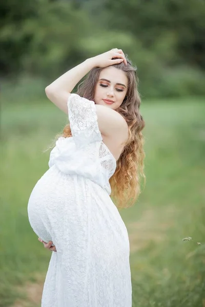 A beautiful young pregnant woman with curly long hair in a white dress walks in the field. Spring portrait of a pregnant woman. Happy pregnancy. Summer.