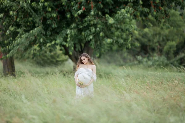 白いドレスを着た中長髪のきれいな若い妊婦さんが畑を歩いています 妊婦の春の肖像画 幸せな妊娠 — ストック写真
