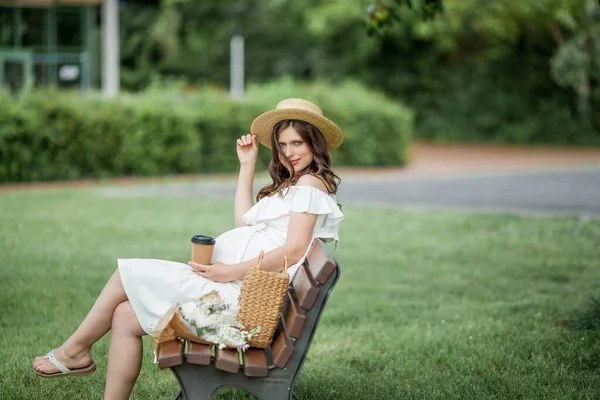 妊娠中の若い美しい女性は公園でカプチーノを飲みます 白いドレスとウィッカーの帽子の妊婦の肖像画 — ストック写真