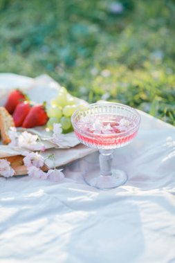 Doğada bahar pikniği. Bir bardak pembe şampanya, sakura çiçekleri, hasır bir çanta, bir şapka, İtalyan..