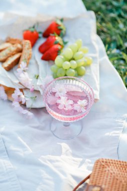 Doğada bahar pikniği. Bir bardak pembe şampanya, sakura çiçekleri, hasır bir çanta, bir şapka, İtalyan..