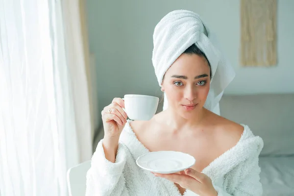 After a shower, a young woman in a bathrobe with a towel on her head drinks coffee and looks out the window. Morning. Breakfast.