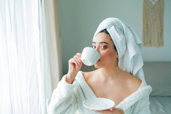После душа молодая женщина в халате с полотенцем на голове пьет кофе и смотрит в окно. Доброе утро. Завтрак.