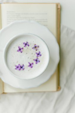 Bej arka planda bir kupa kapuçino. Leylak çiçekleri, kitap. Evde dinlenin. Karantinaya alın. Samimi.