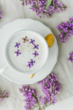 Bej arka planda bir kupa kapuçino. Leylak çiçekleri. Evde dinlenin. Karantinaya alın. Samimi.