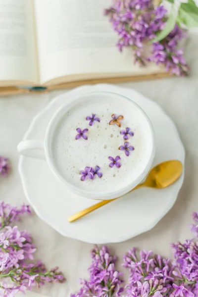 Mug of cappuccino on a beige background. Lilac flowers, book. Rest at home. Quarantine. Cozy.