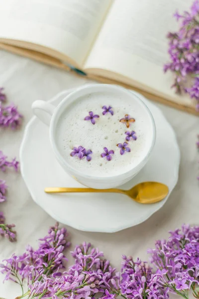 Mug of cappuccino on a beige background. Lilac flowers, book. Rest at home. Quarantine. Cozy.