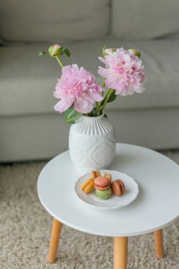 Oturma odasının içi. Vazoda bir buket pembe şakayık, rengarenk Fransız kurabiyesi. Samimi.