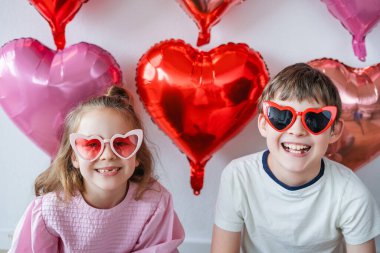 Pembe balonların arka planında kalp gözlüğü takmış şirin erkek ve kız kardeşler. Sevgililer Günü.