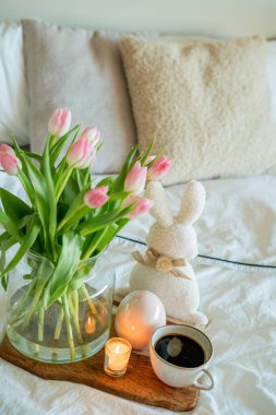 Aydınlık gölgeli bahar Paskalya evi dekorasyonu. Cam vazoda lale buketi, Paskalya tavşanı, tahta tepside sıcak kahve..