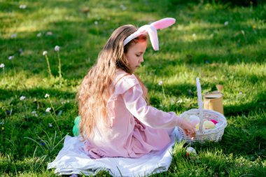 Paskalya havası. Tavşan kulaklı şirin küçük kız çiçek açan bir parktaki çimenlikte renkli yumurtaları topluyor..