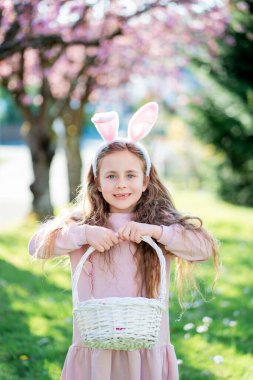 Paskalya havası. Tavşan kulaklı şirin küçük kız çiçek açan bir parktaki çimenlikte renkli yumurtaları topluyor..