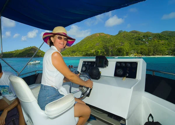 A woman sitting at the helm of a yacht smiles, looking at the photographer. The yacht is approaching the green island. Seychelles.