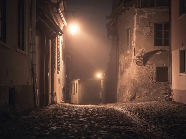 Gece sahnesi Sighisoara Kalesi 'nden, kaldırım taşı sokakları, eski ortaçağ binaları ve lamba direği olan.