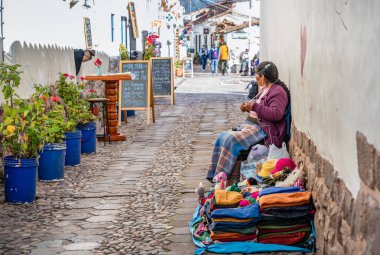 Cusco, Peru - 19 Mart 2025: San Blas Mahallesi 'nde dar, tarihi bir kaldırım taşı caddesi boyunca taştan bir duvarın yanında oturan ve ören yerel esnaf kadın