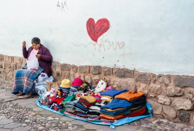 Cusco, Peru - 19 Mart 2025: San Blas Mahallesi 'nde dar, tarihi bir kaldırım taşı caddesi boyunca taştan bir duvarın yanında oturan ve ören yerel esnaf kadın