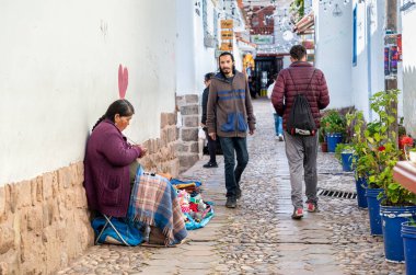 Cusco, Peru - 19 Mart 2025: San Blas Mahallesi 'nde dar, tarihi bir kaldırım taşı caddesi boyunca taştan bir duvarın yanında oturan ve ören yerel esnaf kadın