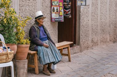 Pisac, Peru - 20 Mart 2025: Pisac kasabasında bir bankta dinlenen ve geleneksel Andean kıyafetleri giyen otantik yerli yaşlı bir kadının portresi.