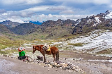 Arka planda Vilcanota sıradağlarının nefes kesici buzul manzarasına sahip bir atın yanında dinlenen geleneksel giysili Perulu kadın.