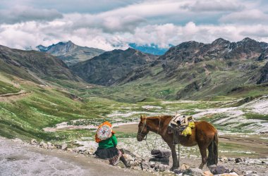 Arka planda Vilcanota sıradağlarının nefes kesici buzul manzarasına sahip bir atın yanında dinlenen geleneksel giysili Perulu kadın.