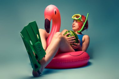 Büyük şişme flamingoda oturan kız, yüzmeye hazır, mavi stüdyo arka planı boş.