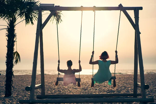 Dos mujeres jóvenes y atractivas balanceándose en el balancín en la playa al atardecer. — Foto de Stock