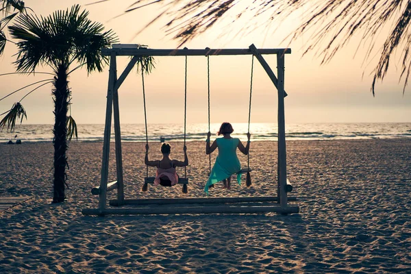 Dos mujeres jóvenes y atractivas balanceándose en el balancín en la playa al atardecer. — Foto de Stock