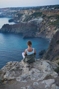 Kadın bir kayanın üzerinde oturuyor, deniz manzarasına, gün batımına bakıyor ve manzaranın ve temiz havanın tadını çıkarıyor..